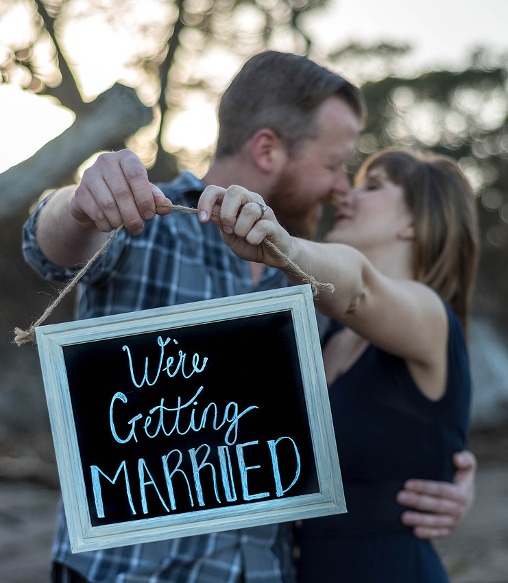 the couple holding a sign saying "We're Getting Married" with the Driftwood trees of Big Talbot Island in the background 